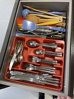 Full view of drawer containing stainless steel cutlery sets separated by type in red plastic tray and wooden and plastic cooking utensils arranged above.