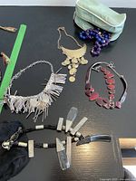 Top view of five costume jewelry necklaces displayed on black surface, showing various styles including beads, metal, fabric fringe, and clear geometric pendants.