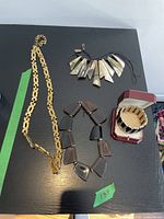 Four jewelry pieces arranged on a black surface: gold-tone chain necklace, chunky dark-tone necklace, natural-tone pendant necklace on cord, gold-tone bracelet in a red box.