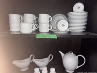 Shelf with stacked white porcelain tea cups, plates, two creamers and two small condiment shakers, and a teapot on lower shelf.