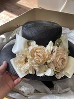 Black woven straw derby hat with wide cream grosgrain ribbon and three large silk flowers in cream and beige.