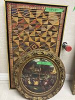 Rectangular framed tapestry or textile art with geometric pattern measuring 28x17 inches leaning against wall with round framed painting in front.