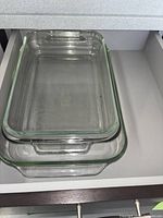 Three clear glass rectangular baking trays stacked inside each other in a drawer. The trays have built-in handles and varying sizes.