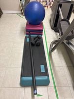 Photo showing aerobic stepper with exercise bar on top, blue stability ball and stack of pink and purple steps in background