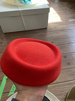 Red handmade hat held in hand, round crown, visible texture of wool felt.