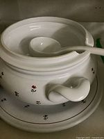 Front view of the white hand painted Italian soup bowl with floral accents, ladle placed inside, and plate underneath