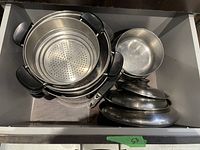 Photo of drawer containing 8 Lagostina stock pots, 2 saucepans without lids, multiple metal lids, and a steamer insert with black handles, all stainless steel.