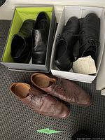 Three pairs of men's leather shoes: two pairs in boxes (black slip-ons and black lace-ups) and one pair brown wingtip shoes outside the box.