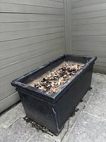 Rectangular black planter pot filled with dry leaves and dirt, showing wear and some discoloration on the outside surface.