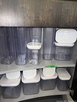 View inside cabinet shelf showing a variety of tall and shorter rectangular clear plastic Oxo storage containers with white lids distributed across two shelves.