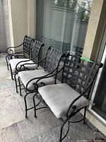 Three black wrought iron patio chairs with lattice backs and light grey cushions arranged outdoors on stone patio.