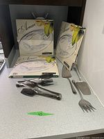 Three boxed Luminarc Arum glass platters and five stainless steel serving utensils displayed on a shelf.