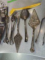 Photo showing assorted silverplated serving cutlery including pie servers, ladle, tongs, and serving scissors laid out on a table.