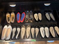 Eleven pairs of ladies ballerina style slip-on shoes arranged in two rows on dark shelves, showing different patterns, colors, and styles.