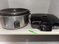 West Bend Crockpot and Hamilton Beach portable grill placed side by side on shelf.