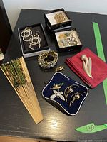 Overview of all items on black tabletop: fan, large earrings, chain necklace, boxed assorted earrings, rhinestone bracelet, zip pouch with brooch, jewelry case with brooches and earrings