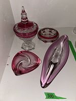 Four pink crystal glass decor items displayed together: covered candy dish, round dish/ashtray, heart-shaped swirl art glass, elongated art glass piece