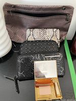 Top view of five wallets stacked together in front of a brown pattern bag and vintage makeup compact open showing powder and brush
