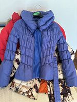 Front view of three women's winter jackets stacked, showing light purple puffer jacket with belt on top, red jacket underneath, and cream floral jacket at bottom