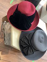 Two derby hats pictured on a wooden floor: a red wool hat with wide black velvet trim and a black derby hat with a suede bow.
