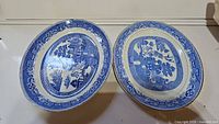 Two vintage blue willow dinner plates side by side on a white surface showing detailed blue and white pattern