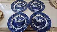 Four blue and white vintage J & G Meakin dinner plates with detailed floral border and central village scene laid out on table.