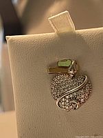 Close-up image of a heart-shaped pendant studded with small clear Swarovski crystals and a polished silver swirl detail, displayed on a cream-colored jewelry stand.