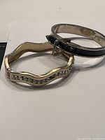 Two bangle bracelets, one gold-tone wavy with clear crystals and one silver-tone with black enamel inset.