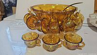Photo showing punch bowl with 10 displayed cups hanging on its side rim and 3 cups in front, highlighting the iridescent amber carnival glass and grapevine pattern.