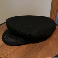 Side and front angled view of the black traditional Greek fisherman's cap, placed on wooden surface, showing embossed pattern on visor and felt material texture.