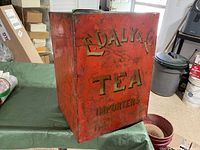 Front side of tall rectangular red metal tea tin with yellow stenciled 'E. Daly & Co Tea Importers' text, showing surface wear and scratches.