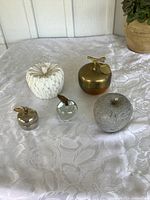 Clear image showing all five decorative apple items on a white textured cloth background: white decorative apple, large brass apple trinket box with lid and bird detail, clear blown glass paperweight apple with brass leaf and stem, silver small trinket box apple, and gray stone-like decorative apple.