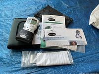 Photo showing the complete lot including the Aura Wave Perfect T.E.N.S. device in its black carrying pouch, user manual, a sealed adhesive electrode pad pair, and additional instructional paper.