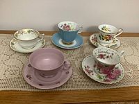 Five vintage china cups and saucers displayed on lace runner with distinct floral and gold trimmed designs, showing variety and condition.