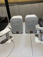 Pair of white JBL Control 1 Pro-5 compact speakers displayed side by side on a table with two white metal speaker mounts.