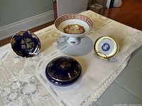 Four Limoges porcelain items displayed on lace tablecloth including pedestal dish, covered jar, and two decorative plates one on stand.