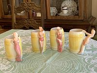 Four barrel-shaped mugs with yellow glaze and lady figurine handles in various poses set on table.