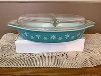 Side view of turquoise Pyrex casserole dish with snowflake design and clear divided glass lid, on white box and lace runner.