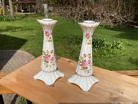 Pair of white china candlesticks with pink floral designs and gold edges placed on wooden table outside in sunlight with garden background.