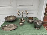 Overall view of silverplate set showing raised Sheffield plate, oval tray, creamer, sugar bowl, Viking rose bowl with perforated lid, and folding candelabra on green cloth in front of garage door.