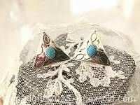 Pair of triangular sterling silver earrings placed on lace fabric backdrop, showing front side with turquoise stones and etched silver design.