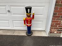Full front view of 40 inch tall wooden toy soldier figure standing outdoors on black round base in front of white garage door.