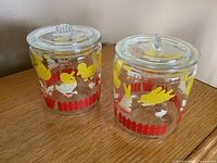 Two clear glass jars with lids featuring yellow farm animal stencils, red picket fence, and flowers on wooden surface.