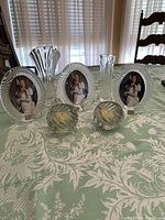 Group shot of all crystal items on a patterned tablecloth including 3 Mikasa oval photo frames, 2 small round swirl candleholders, Mikasa Flores bud vase and D’Arques bud vase.
