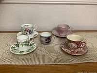Full view of 5 demitasse cups and saucers laid on a lace table runner showing front and top views highlighting patterns and colors.