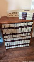Full view of wooden shelf housing 27 volumes including 23 volume Encyclopedia Britannica set, other volumes, and box labeled Britannic World Atlas on top