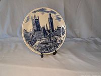 Photo of the front of a blue and white ceramic plate showing a painted and engraved imagery of Big Ben, river boats, and surrounding architecture on a flat blue sky background.