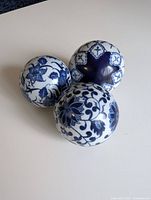 Photo showing three blue and white painted glass balls with floral and geometric decorative patterns, arranged closely on a white surface.