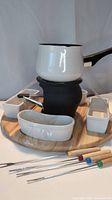 Photo of the complete fondue set showing the white metal pot on the black metal base, four white side containers on wooden base, and four fondue forks