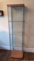 Full view of the tall glass display case against a wall, showing its wooden top and base with glass panels and two shelves inside.